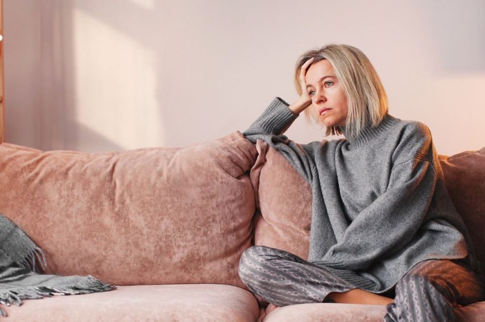 Mulher na casa dos quarenta sentada no sofá, pensativa e cansada, a refletir sobre as mudanças emocionais e energéticas desta fase de transição.