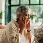 Mulher a sentir um afrontamento durante a menopausa, refrescando-se em frente a uma ventoinha, representando a busca de alívio natural e consciente.