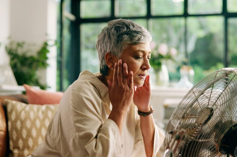 Mulher a sentir um afrontamento durante a menopausa, refrescando-se em frente a uma ventoinha, representando a busca de alívio natural e consciente.
